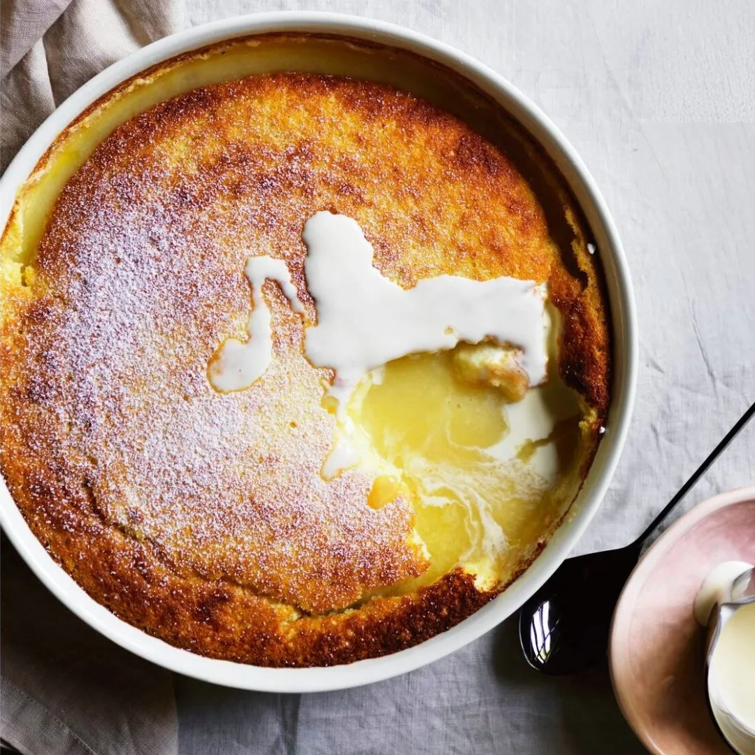 Over-the-top shot of a round white dish with a baked lemon pudding, with a spoonful taken out of the crust to reveal the molten lemon curd inside.