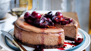 photo of table with white tablecloth and a blue-rimmed plate underneath a brown cheesecake topped with glossy cherries