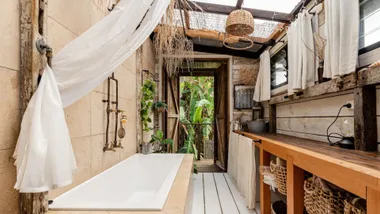 Rustic bathroom with a white bathtub, wooden counter, woven baskets, and greenery outside the open door.