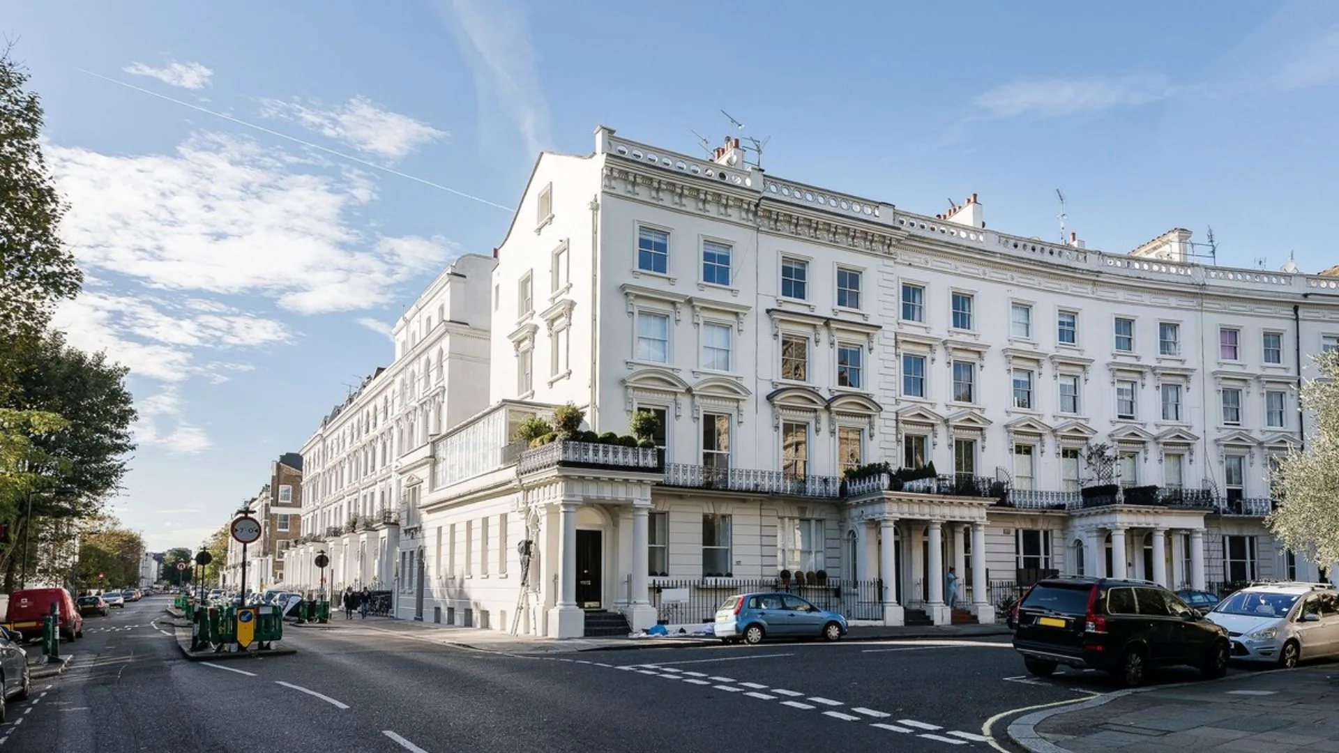 Street view of elegant white terraced Victorian townhouses with parked cars and a blue sky.