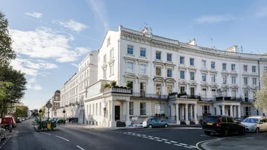 Street view of elegant white terraced Victorian townhouses with parked cars and a blue sky.