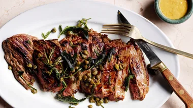 Grilled steak with herbs and capers on a white plate, accompanied by a fork and knife, with a bowl of mustard sauce.