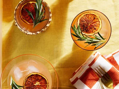 Three burnt grapefruit and rosemary palomas in glasses with sprigs of rosemary, gold fork and white and orange check napkin.