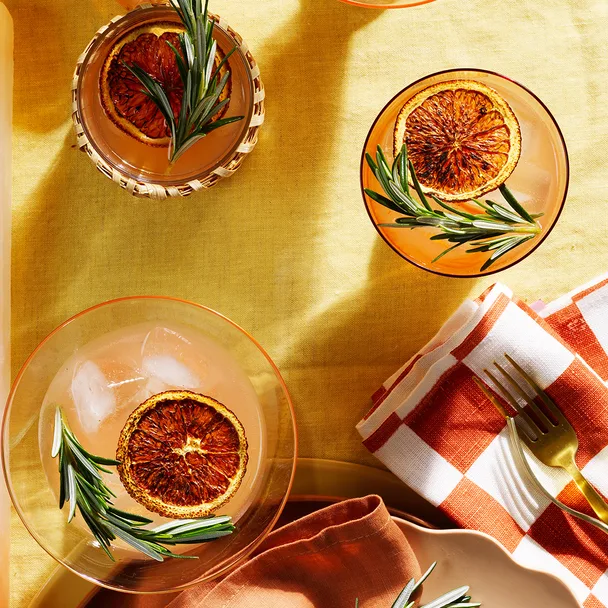 Three burnt grapefruit and rosemary palomas in glasses with sprigs of rosemary, gold fork and white and orange check napkin.