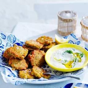 Chickpea fritters in light blue dish with blue and white paper and herb and garlic yoghurt in small yellow dish to side.