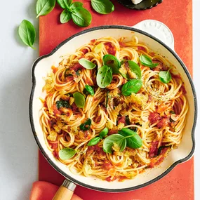 Crunchy garlic and tomato pasta on white and blue cooking pan with scattered basil leaves.