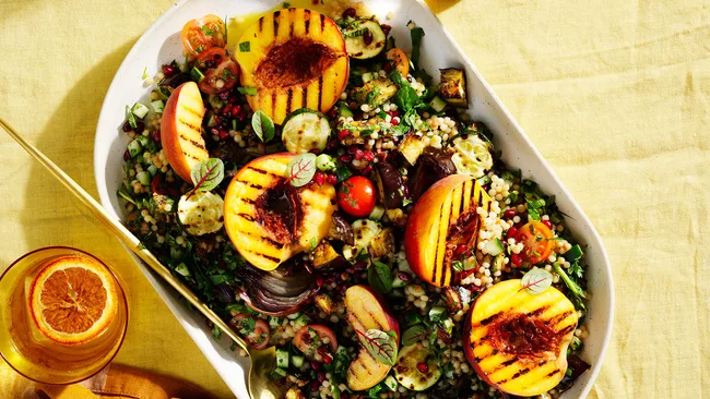 Eggplant with tabbouleh salad with grilled peaches and pomegranate on white dish with gold spoon and small bowl of tabbouleh salad.