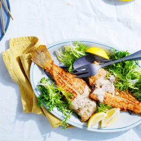 Flounder with avgolemono sauce with salad and lemon on light blue dish with grey cutlery.