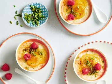 Greek rice puddings with pistachio, lemon and fresh raspberries on white and orange rimmed plates with white spoons and small blue bowl of pistachios.