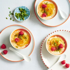 Greek rice puddings with pistachio, lemon and fresh raspberries on white and orange rimmed plates with white spoons and small blue bowl of pistachios.