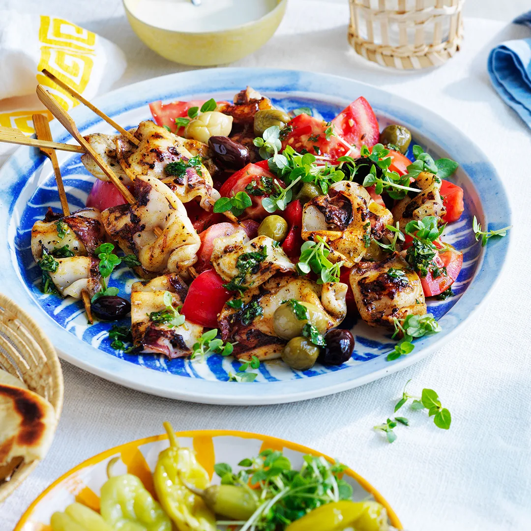 Grilled calamari salad with yoghurt and ouzo dressing on blue serving platter with pickled peppers in orange and white dish to side.