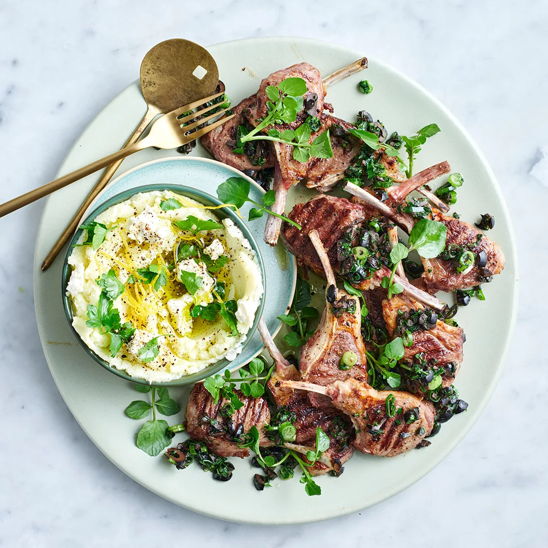 Lamb cutlets with feta skordalia and micro herbs on white plate with gold spoon and fork.