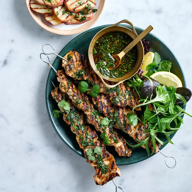 Pork skewers with chimichurri, salad and lemon in dark green bowl alongside grilled potatoes in a pink bowl.