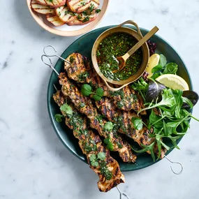 Pork skewers with chimichurri, salad and lemon in dark green bowl alongside grilled potatoes in a pink bowl.