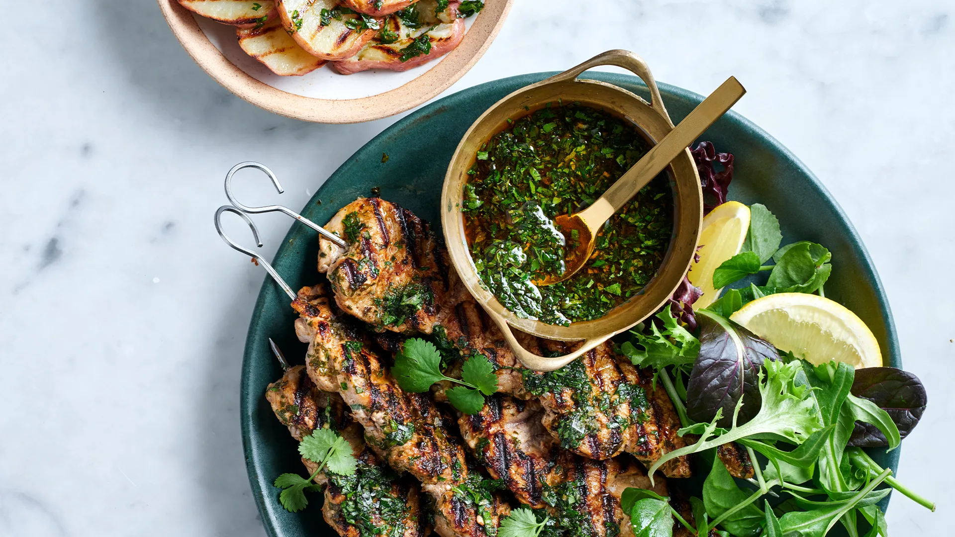 Pork skewers with chimichurri with salad and lemon in a dark green bowl with grilled potatoes in a pink bowl.