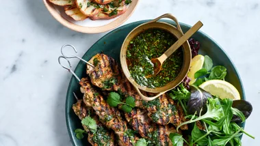 Pork skewers with chimichurri with salad and lemon in a dark green bowl with grilled potatoes in a pink bowl.