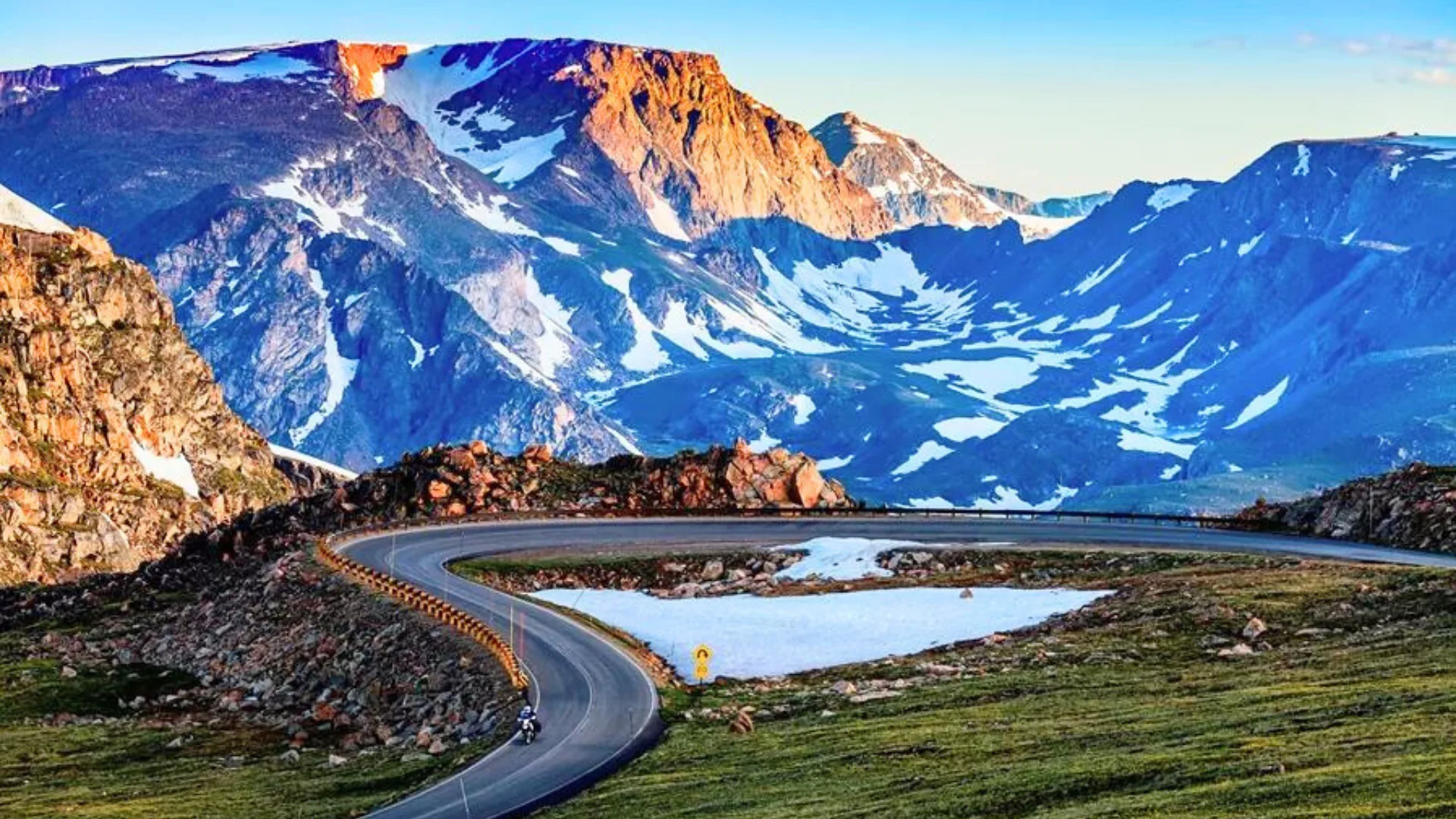 Mountain road with a sharp curve, surrounded by rocky peaks and patches of snow, under a clear blue sky.