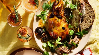 Slow-cooked lamb shoulder with black garlic and mint persillade on pink dish with white and orange check napkin and two grapefruit palomas with burnt grapefruit and a sprig of rosemary.