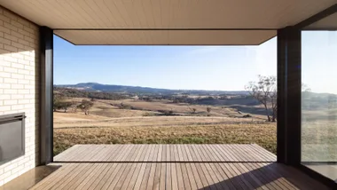 View from a modern room with wood flooring, showcasing a vast landscape of hills and clear blue sky through large windows.
