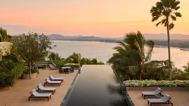 Amanpuri Phuket resort infinity pool overlooking a bay at sunset, with white sun lounges on either side of pool