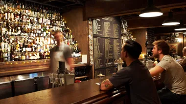 Photo of two men sitting at the bar and bartender blurred in motion in front of large whisky collection in back bar at Baxter Inn in Sydney, which is one of the best bars Australia that have had a lasting impact