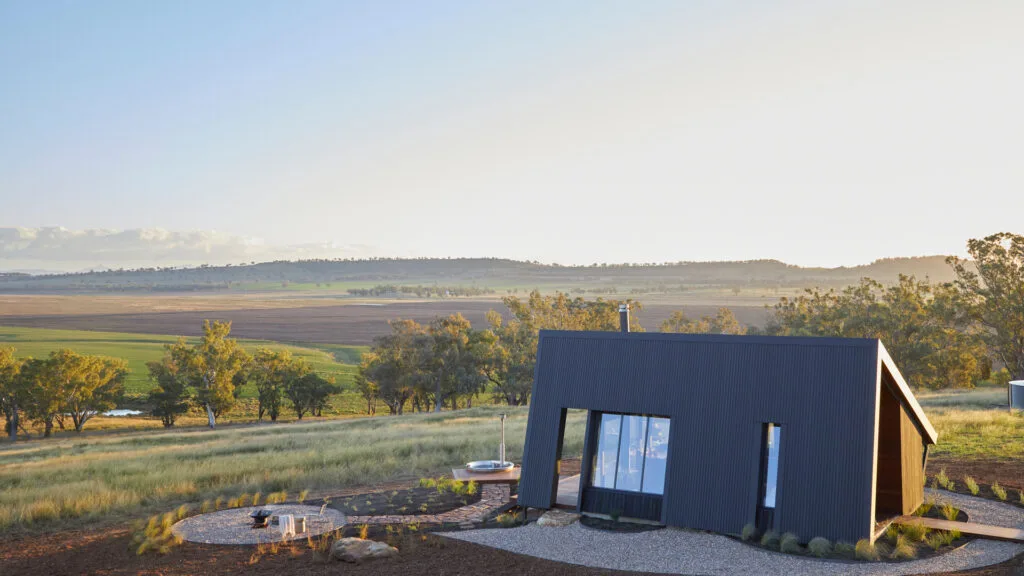 Tiny home NSW stay Gilay Estate in Quirindi, showing the black cabin exterior in daylight with neat landscaped garden, fire pit and outdoor hot tub with views over the countryside