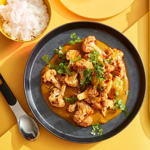 Korean cauliflower curry on black bowl with a bowl of rice and spoon to the side on a yellow background.