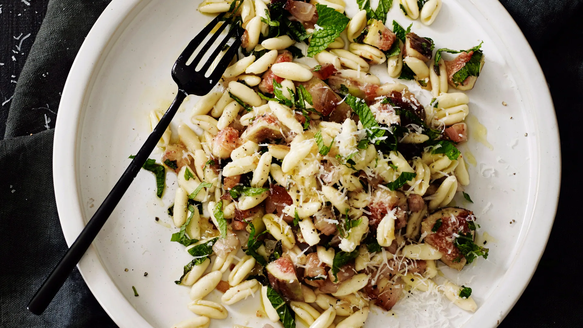 Cavatelli pasta shapes with pancetta, mint, figs and pecorino recipe on white plate with black long fork and dark coloured background