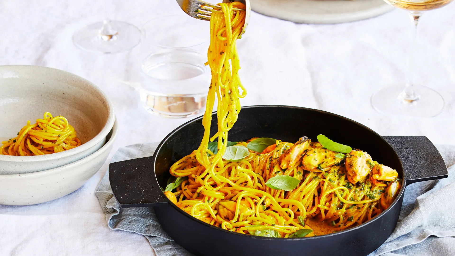 Pasta types: Fork holding up ribbons of saffron tagliolini pasta with mussels, lemon and basil from a black bowl with white table setting background