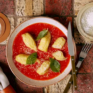 Bowl of culurgiones on a bed of passata garnished with basil on wood background. Bowl of pecorino to the side.