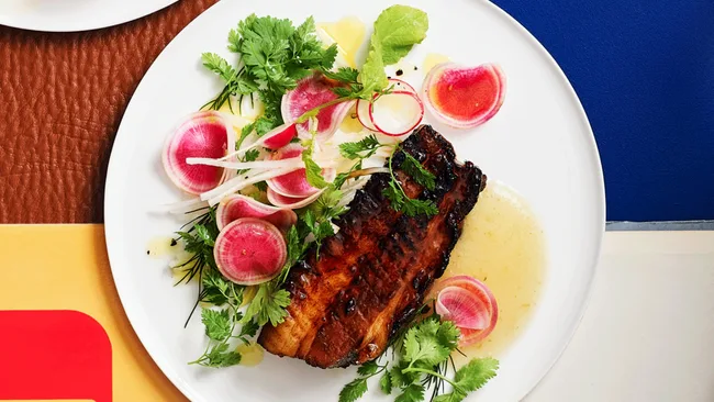 Grilled meat with salad including radishes, herbs, and dressing on a white plate with a blue and brown background.