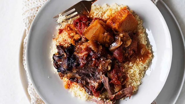A plate of couscous topped with braised lamb, tomatoes, and spices, with a fork on the side.