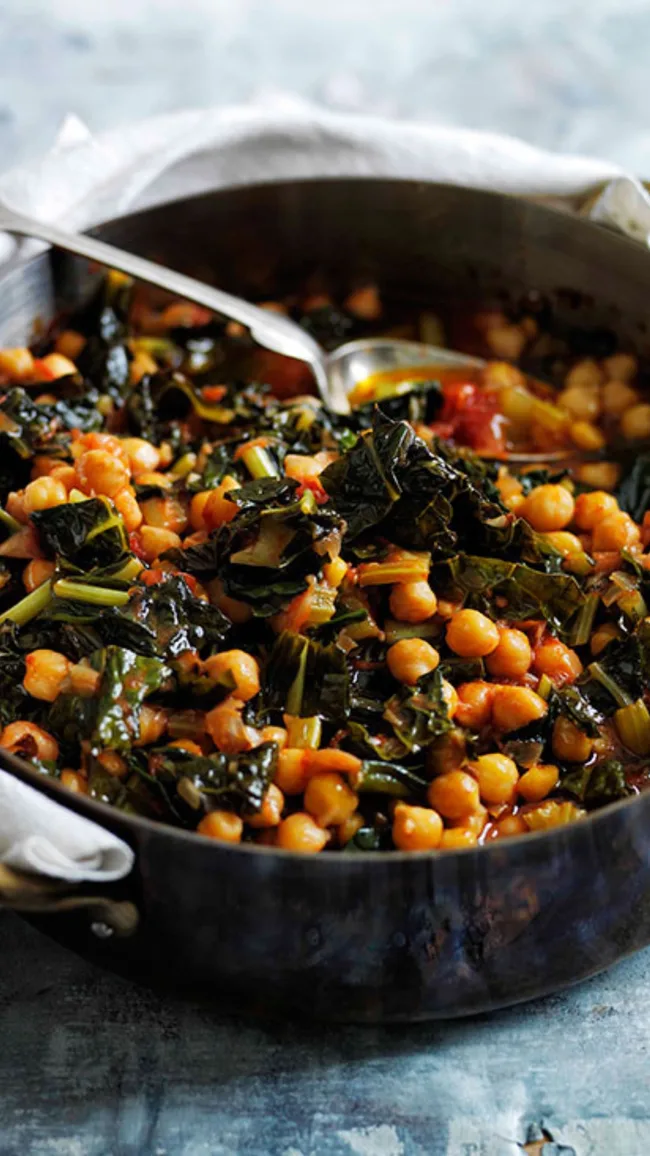 A pot of chickpeas and kale stew with a spoon, placed on a textured blue surface.