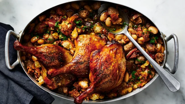 Roasted duck legs on a bed of beans, sausage, and herbs in an oval dish, with a serving spoon on the side.