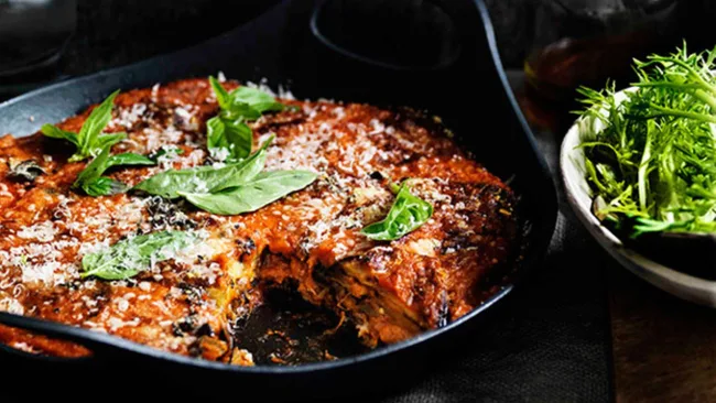 Baked lasagna topped with basil leaves in a black dish, accompanied by a bowl of green salad.