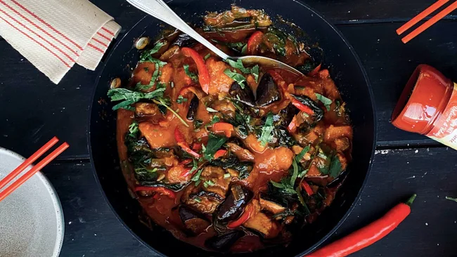 A vibrant Thai red curry dish in a black bowl, garnished with herbs and served with chopsticks on a dark wooden table.