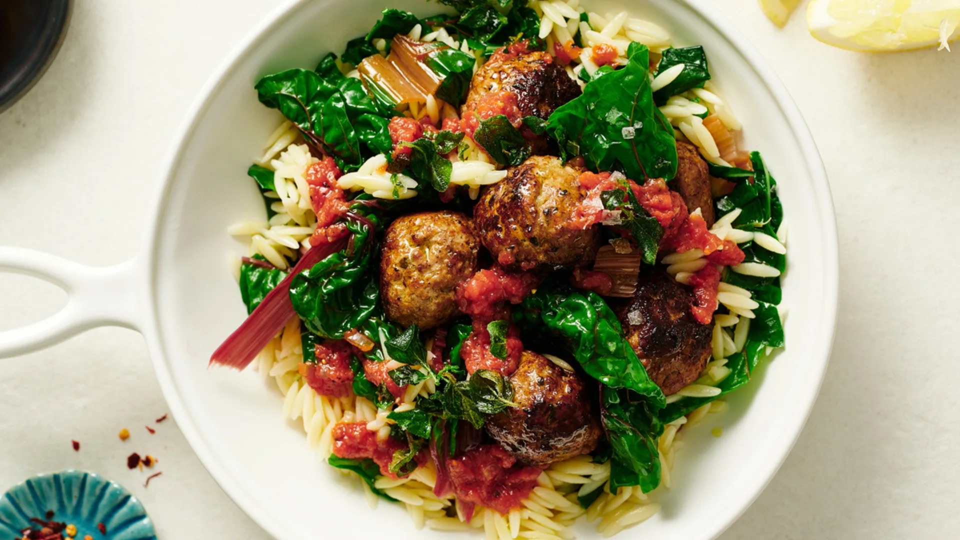 A white bowl of orzo pasta with meatballs, sautéed greens, and tomato sauce.