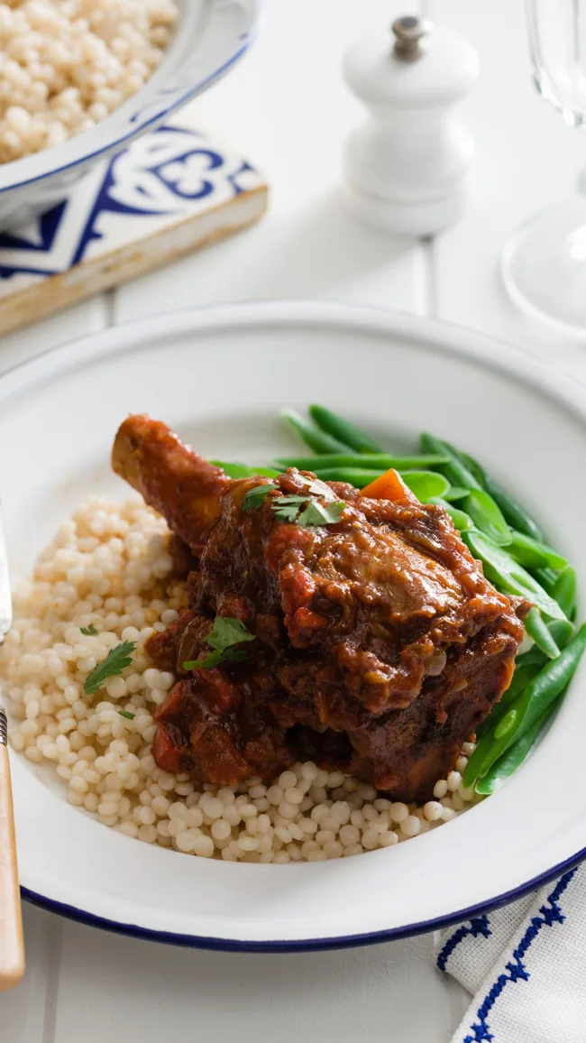 Braised lamb shank with tomato sauce served on couscous, accompanied by green beans on a white plate.