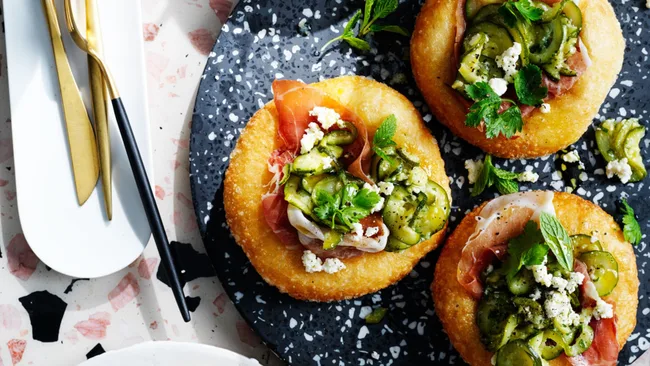 Three fried dough disks topped with prosciutto, sliced cucumbers, feta, and fresh herbs on a black speckled plate.
