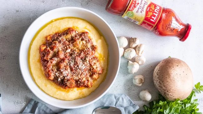 Bowl of polenta topped with tomato sauce, surrounded by mushrooms, parsley, and a bottle of Mutti tomato puree.