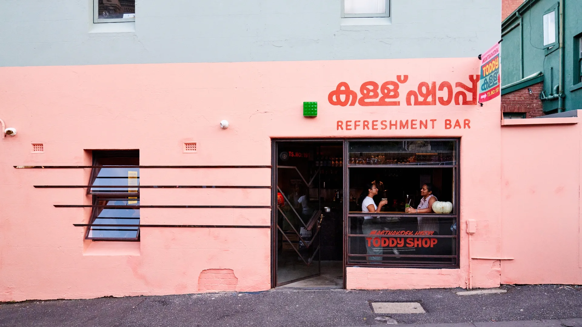 The exterior of a restaurant with a pink wall. Two people are sitting in the window.