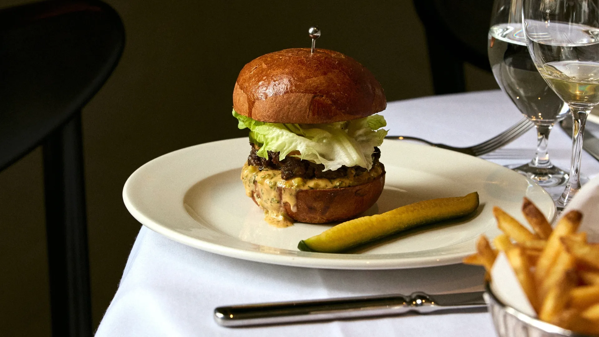 A burger on a table with a white tablecloth, with chips and a glass of wine next to it.