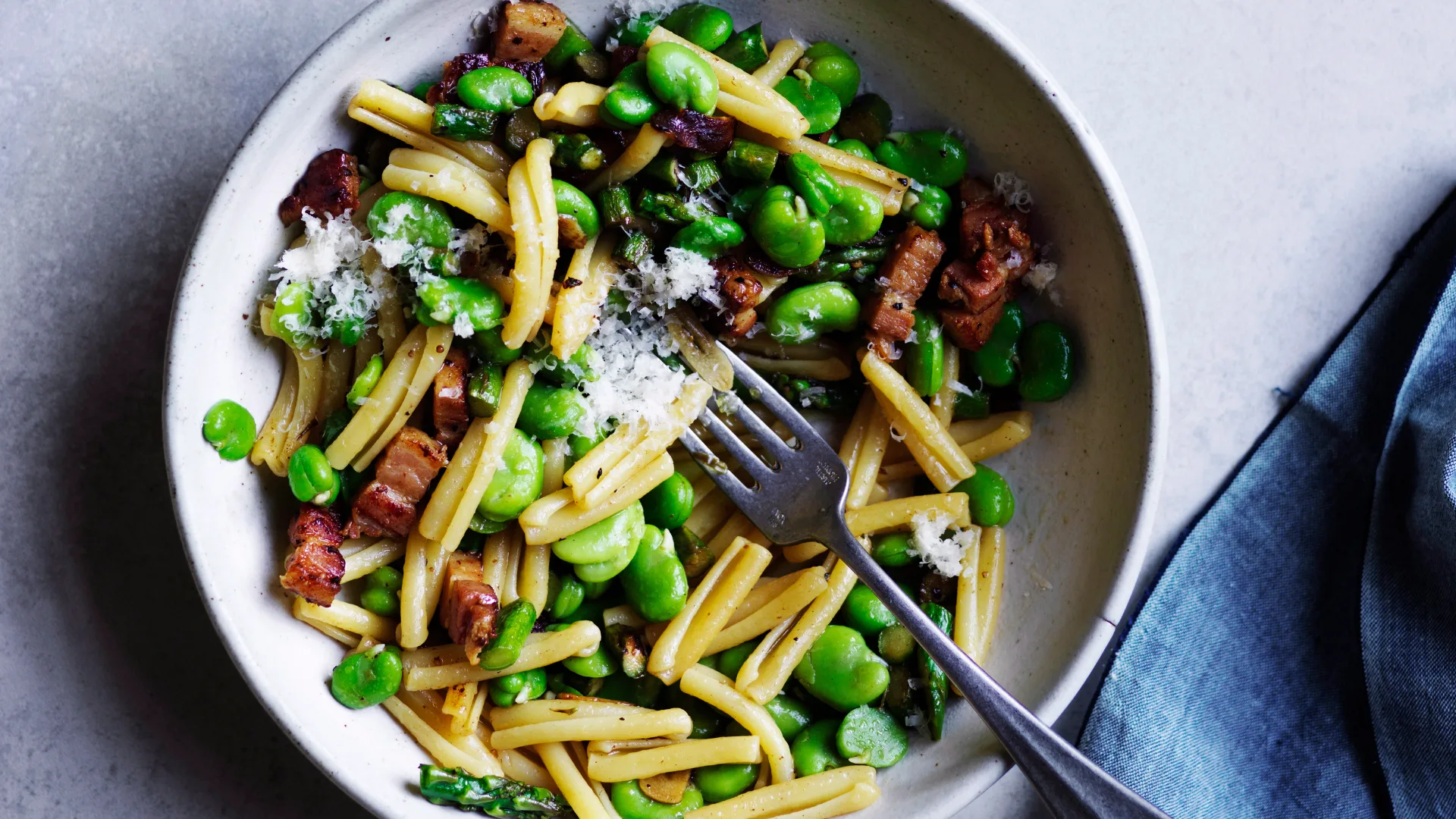 White bowl holding a mixture of short pasta, broad beans, asparagus and pancetta, with a steel fork inside, and a blue napkin to the side.