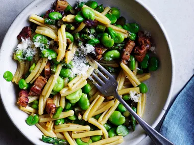 White bowl holding a mixture of short pasta, broad beans, asparagus and pancetta, with a steel fork inside, and a blue napkin to the side.