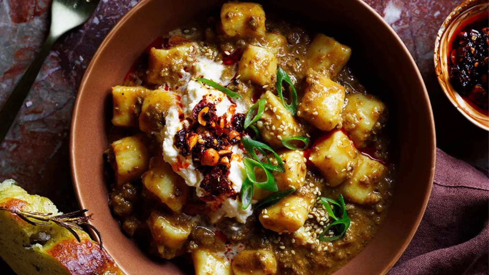 An overhead shot of a bowl of ricotta gnocchi in a mapo-inspired bolognese sauce with cutlery and charred bread