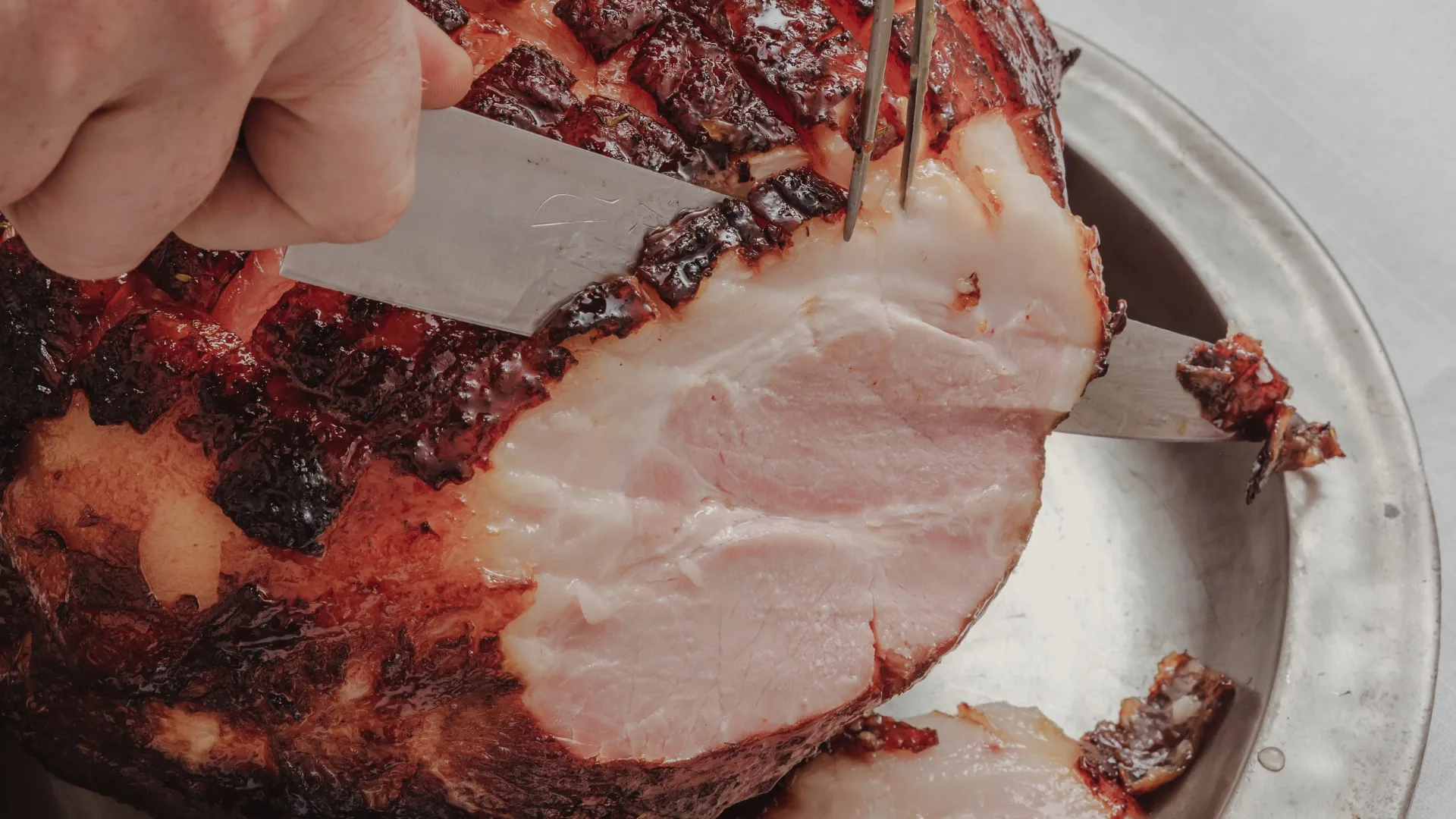A close-up shot of a scored and roasted ham being cut with a knife to emit a pink flesh with a layer of fat