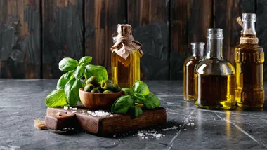 wine, olives and basil leaves on a square wooden chopping board, with 3 more pints of olive oil on the right