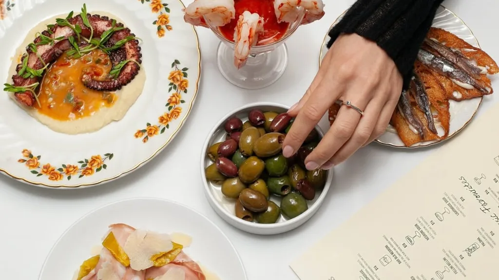 An overview show of a hand reaching for a plate of olives, besides a cocktail glass of cooked prawns and plate of octopus for Bar Olo's happy hour.