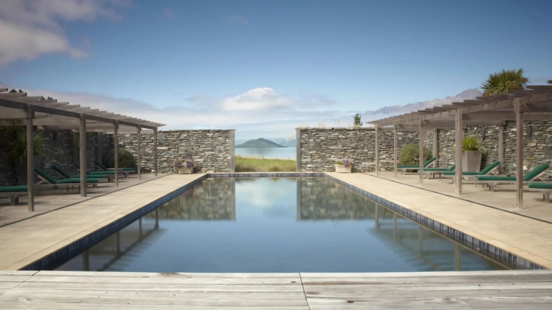 Outdoor pool with lounge chairs and stone walls, overlooking a scenic mountain and lake view under a clear blue sky.