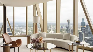 Modern living room with large windows, city view, beige curved sofa, glass coffee tables, and a brown chair.